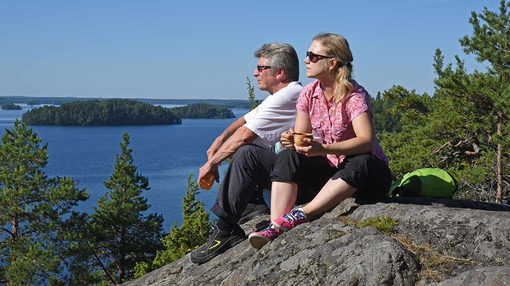 Two hikers are sitting on a cliff. Lake scenery in the background.