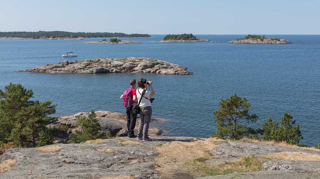 Two people look out to sea. There are rock formations and a sailboat in the background.
